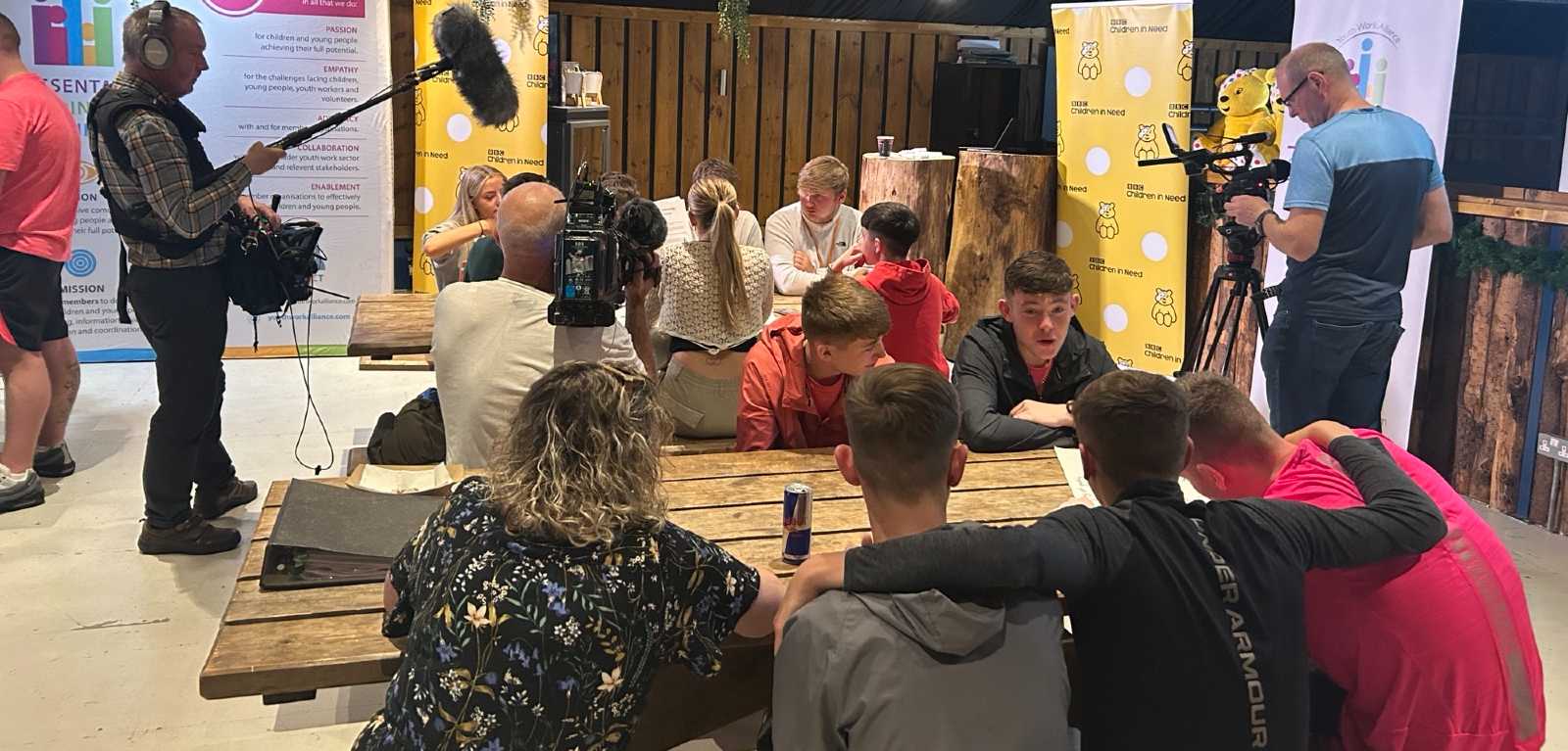 teenage boys and girls sitting at two wooden rectangular tables with camera and sound staff beside them